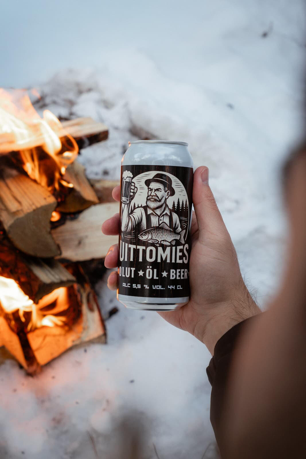 Uittomies olut henkilön kädessä. Taustalla nuotio talvisessa maastossa.