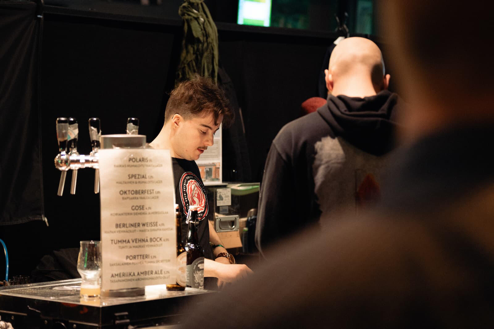 Panimon työntekijä palvelee asiakasta tiskin takana Lakeuden Panimojuhlissa, kuvattuna toisen ihmisen olan takaa.