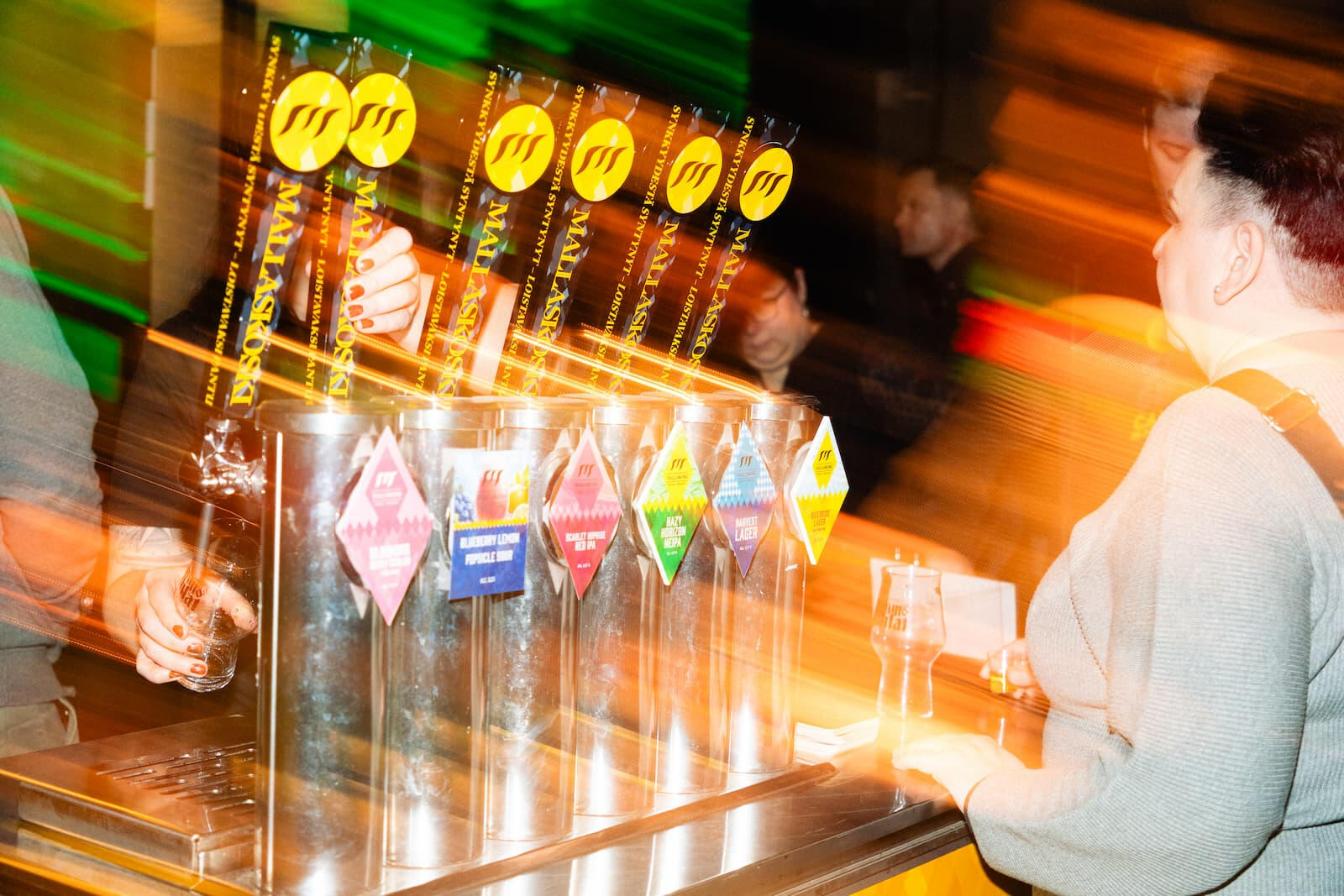 Värikkäät oluthanat Mallaskosken toimipisteellä, olutta kaadetaan hanasta ja asiakas odottaa, kuvattu liike-epäterävyydellä.