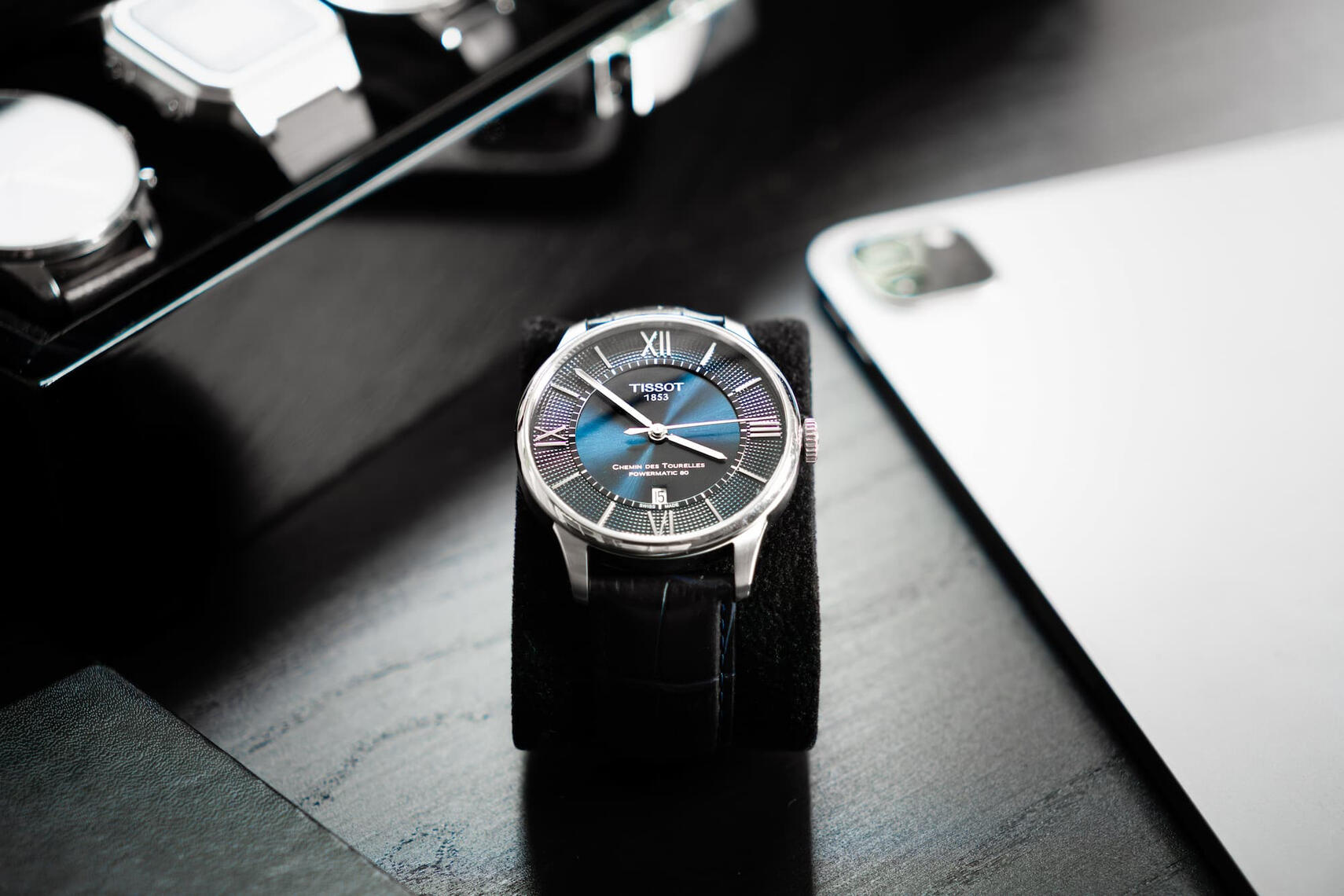 Lähikuva Tissot Chemin des Tourelles -rannekellosta pöydällä, mustassa nahkarannekkeessa, valaistuna pehmeällä luonnonvalolla