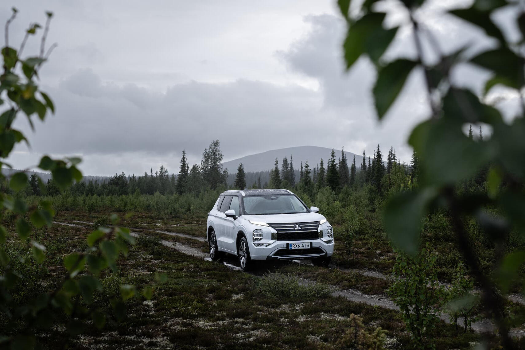 Mitsubishi Outlander etuviistosta kuvattuna sateisella metsäaukiolla, taustalla tunturi.