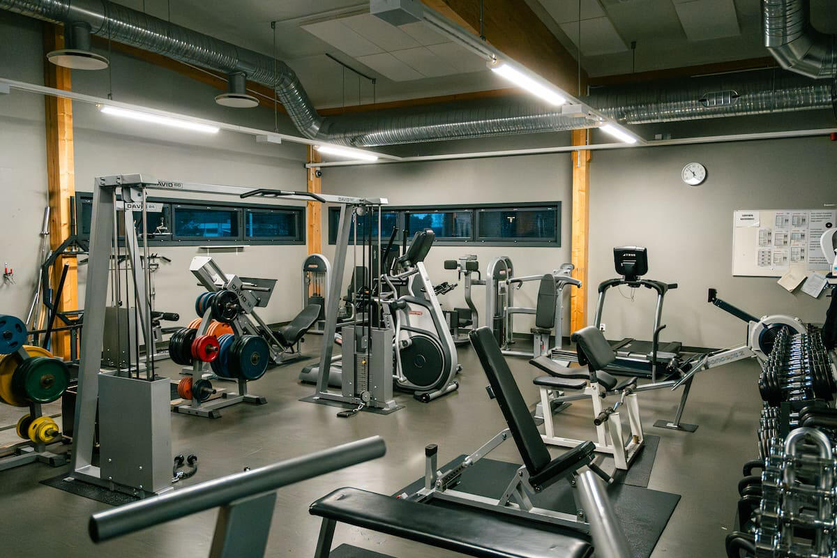 Sisäkuva kuntosalista, jossa näkyy kuntolaitteita ja painoja. Valo tulee kattovalaisimista ja kuvassa korostuu tilan selkeys ja valaistuksen tasaisuus.