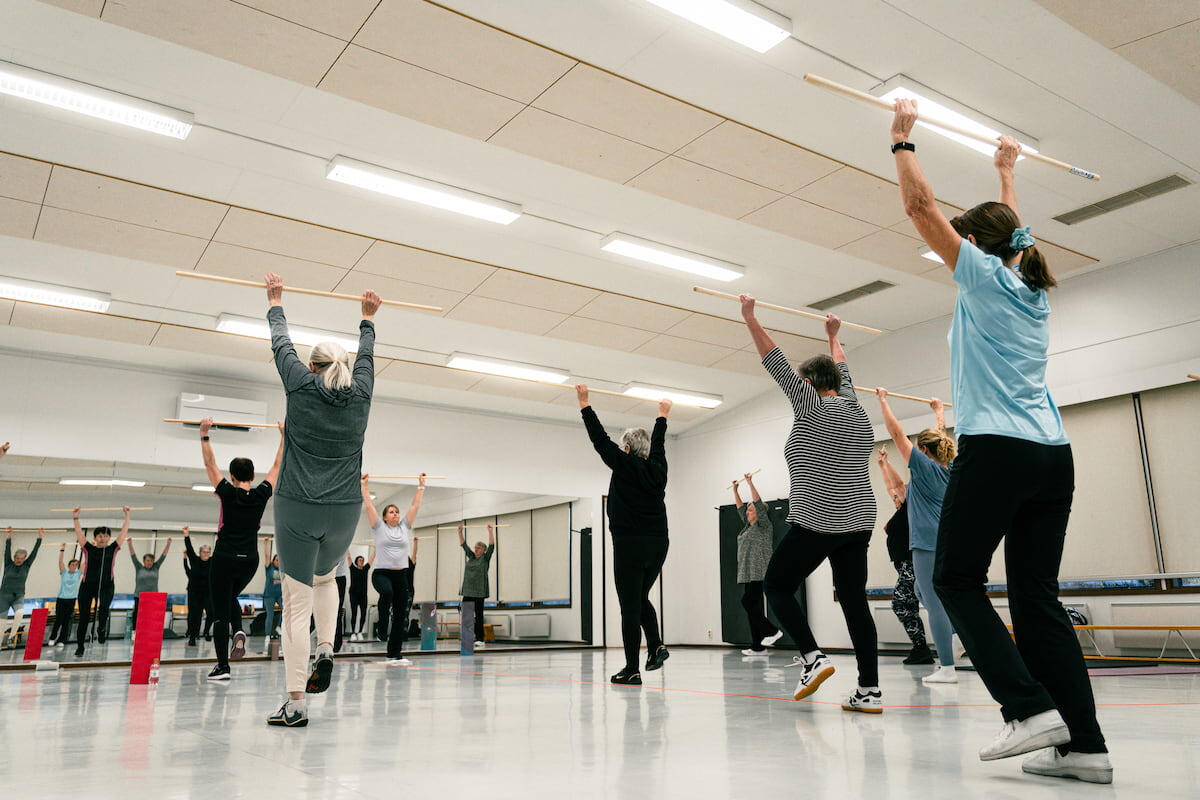 Ryhmä ihmisiä harjoittelee sisäliikuntatilassa kepit käsissä, heijastuen taustalla näkyvistä peileistä. Kuva on otettu matalalta kulmalta korostaen liikkeen dynaamisuutta.